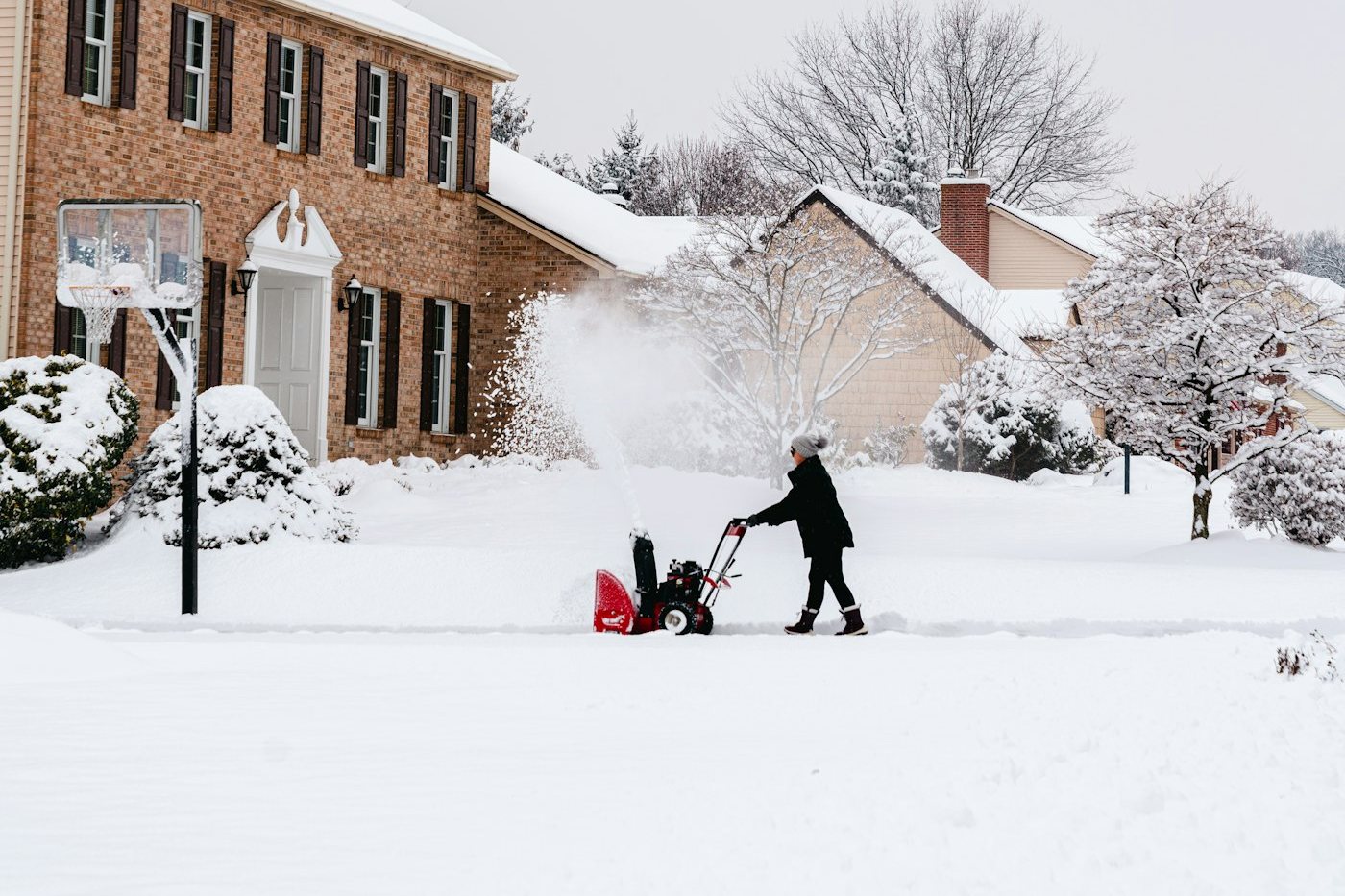 A person clearing a snowy driveway with a red snow blower in front of a brick house