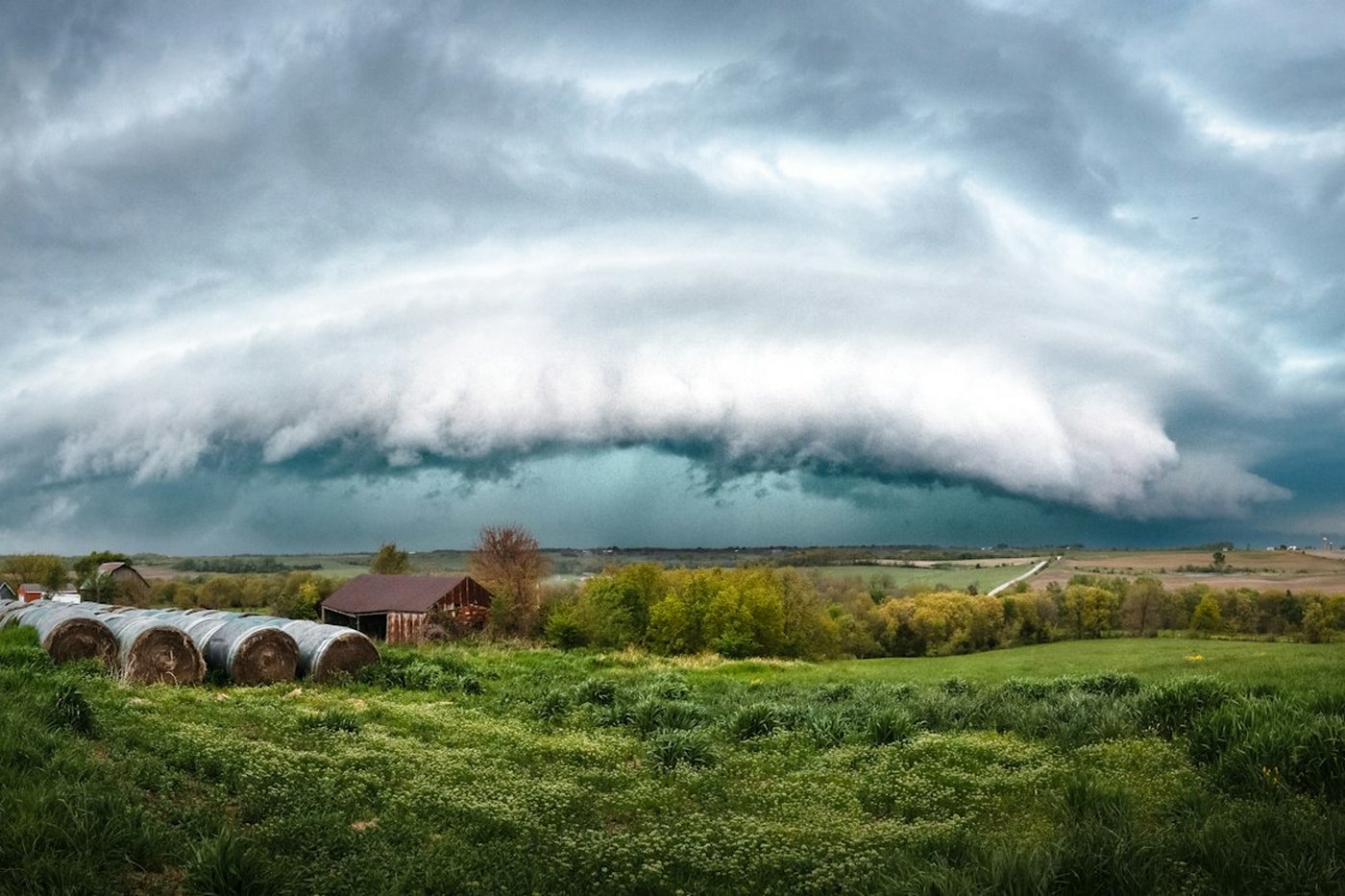 A huge shelf cloud rolling over green farmland and hay bales ahead of a summer thunderstorm